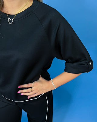 A person wearing a black oversized short-sleeve top with rolled cuffs and black pants with white piping