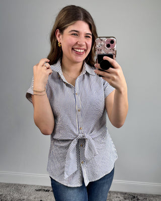 Woman taking a selfie wearing a striped shirt with a tie waist against a plain background