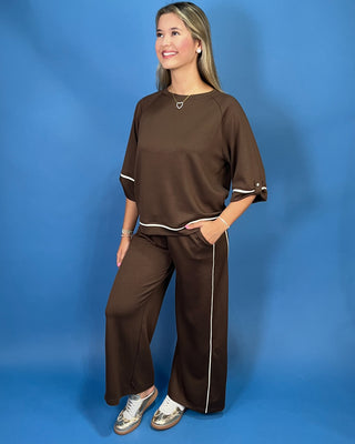 A woman modeling a matching brown lounge set with white piping details, consisting of a short-sleeve top and wide-leg pants