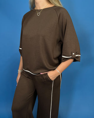 A woman wearing a brown two-piece lounge set consisting of a short-sleeve top with rolled cuffs and matching wide-leg pants with white piping