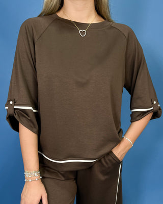 A woman wearing a brown long-sleeve top with white contrast trim on the sleeve cuffs and hem, paired with matching brown pants