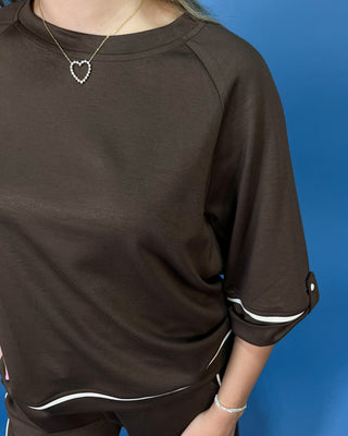 A person wearing a dark brown short-sleeve top with white contrast trim along the hem and rolled cuffs, paired with matching dark bottoms