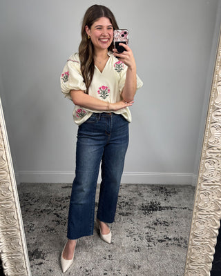 Woman taking a mirror selfie wearing an embroidered blouse and blue jeans.