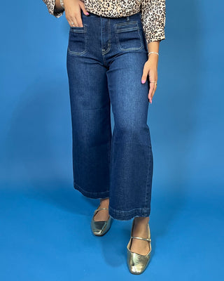 A person wearing dark blue wide-leg cropped jeans, a leopard print top, and metallic gold Mary Jane shoes