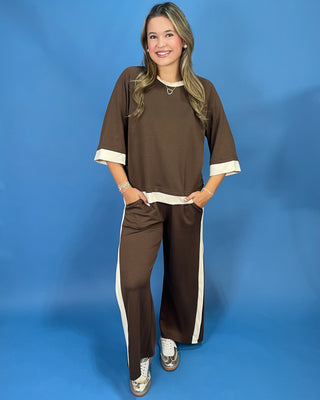 A woman modeling a matching brown two-piece lounge set with cream-colored trim, consisting of a short-sleeve top and wide-leg pants, set against a solid blue studio backdrop.