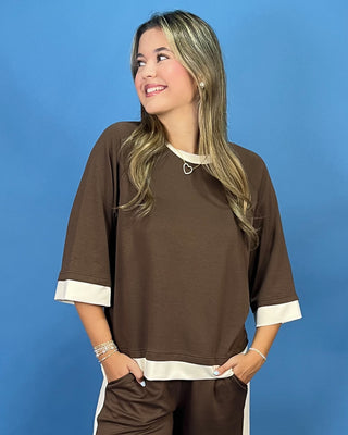 A woman wearing a brown short-sleeve top with cream cuffed sleeves and hem, paired with matching brown pants with cream side stripes, posed against a solid blue background.