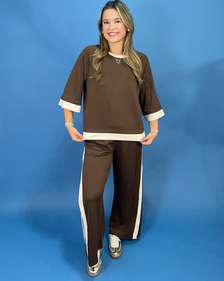 A woman modeling a matching brown lounge set with cream trim, consisting of a short-sleeve top and wide-leg pants, against a solid blue studio backdrop.