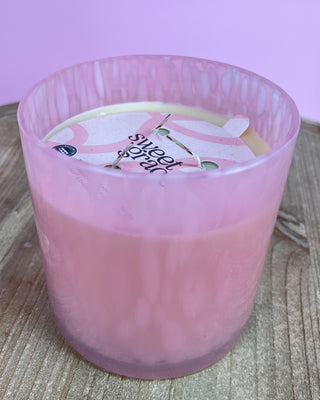 Pink candle in a glass container with a visible brand label on a wooden surface.