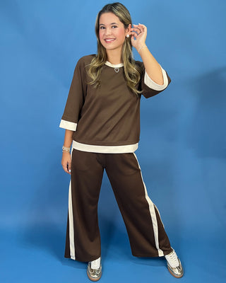 A woman modeling a matching brown two-piece lounge set with cream trim, consisting of a short-sleeve top and wide-leg pants, against a solid blue studio background.