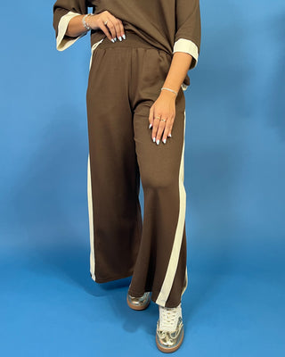 A person wearing brown wide-leg pants with cream side stripes and a matching brown top with cream cuffs, standing against a solid blue background.