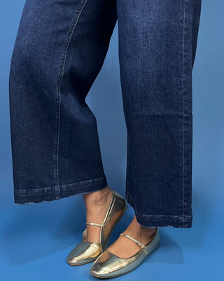 A close-up of a person's legs wearing dark blue wide-leg jeans and metallic gold Mary Jane flat