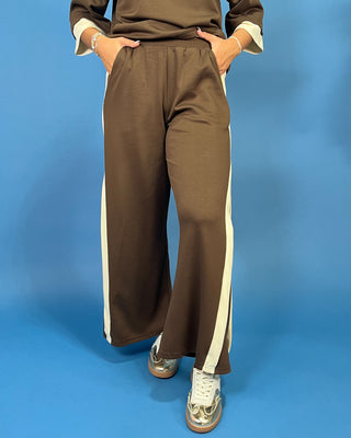 A person wearing brown wide-leg track pants with white side stripes, paired with a matching brown top and white sneakers, standing against a solid blue background.