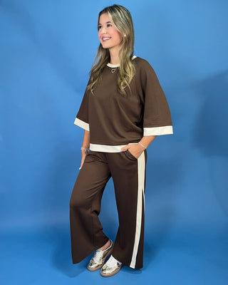 A woman modeling a brown two-piece lounge set with white contrast trim, featuring a short-sleeve top and matching wide-leg pants, posed against a solid blue studio backdrop.