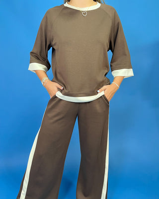 A person wearing a brown two-piece lounge set with white contrast trim, featuring a short-sleeve top and matching wide-leg pants, posed against a solid blue background.