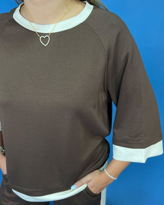 A person wearing a brown raglan-style top with white contrast trim at the crew neck, sleeve cuffs, and hem, photographed against a bright blue background.