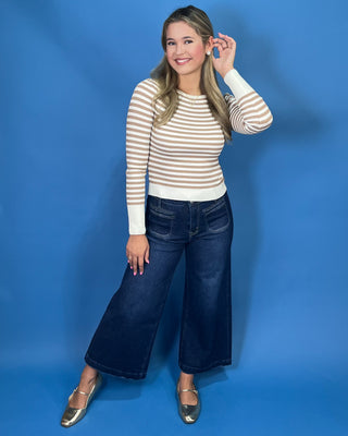 A woman modeling a beige and white horizontally striped long-sleeve knit top with dark wide-leg jeans and metallic gold flats