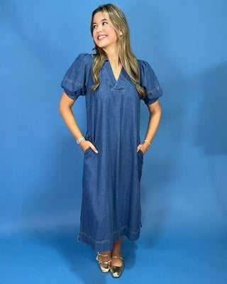 A woman modeling a short-sleeve denim maxi dress with pockets, standing against a solid blue studio backdrop.