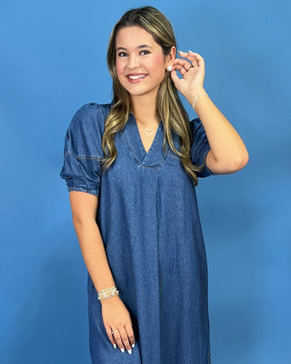 A woman with blonde-highlighted hair wearing a short-sleeve blue denim dress, posing against a solid blue studio background.