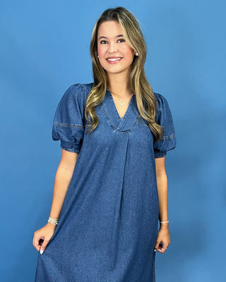 A woman modeling a blue denim dress with short puff sleeves and a V-neckline, standing against a solid blue background.