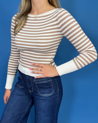 A woman wearing a beige and white horizontally striped ribbed knit sweater with a boat neckline, paired with dark blue jeans