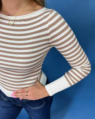 A close-up of a woman wearing a ribbed long-sleeve knit top with horizontal brown and off-white stripes, paired with blue jeans