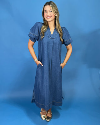A woman modeling a blue denim midi dress with puff sleeves, a V-neckline, and side pockets, standing against a solid blue studio backdrop.