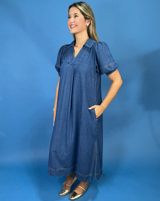 A woman modeling a short-sleeve blue denim midi dress with a V-neck and pockets, standing against a solid blue studio backdrop.