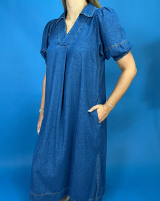 A woman wearing a blue denim midi dress with puff sleeves, a V-neckline, and side pockets, standing against a solid blue studio backdrop.