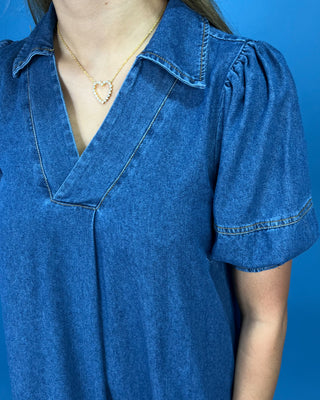 A close-up of a person wearing a blue denim short-sleeve top with a V-neck and puff sleeves, paired with a delicate gold heart necklace, against a solid blue background.