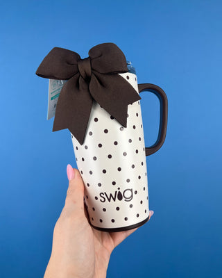 A hand with pink nail polish holds a white insulated tumbler with black polka dots and a large dark brown bow tied around the handle. The tumbler has the brand name 'swig' printed near the bottom, set against a solid blue background.