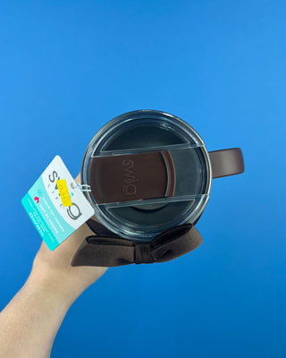 A hand holding a dark brown insulated travel tumbler with a clear lid, brown silicone straw cover, and a teal and white 'STOCK' hangtag against a solid blue background.