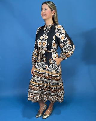 A woman modeling a long-sleeved midi dress with a black base and intricate white, gold, and brown bohemian-style floral and geometric patterns, standing against a solid blue studio backdrop.
