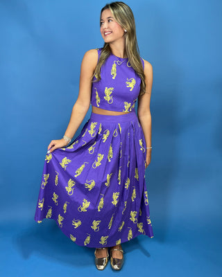 A woman modeling a purple sleeveless two-piece set consisting of a crop top and a flared midi skirt, both printed with a yellow lion pattern, set against a solid blue studio background.