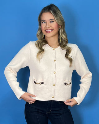 Ivory knit cardigan with gold buttons, long sleeves, front pockets, and decorative gold hardware.
