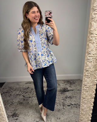 Woman taking a mirror selfie wearing a floral blouse and jeans, standing in front of a decorative mirror.