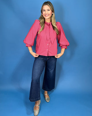 A woman modeling a pink and white vertically striped long-sleeve blouse with a tie-neck detail, paired with dark blue cropped jeans and silver shoes
