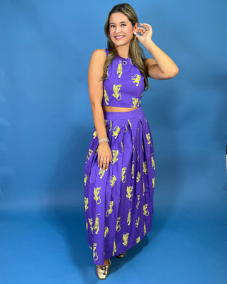 A woman modeling a purple sleeveless two-piece set with a matching crop top and pleated maxi skirt, both printed with a yellow seahorse pattern, set against a solid blue studio backdrop.