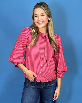 A woman modeling a pink and white vertically striped long-sleeve blouse with a tie-neck detail, paired with dark blue cropped jeans and silver shoes