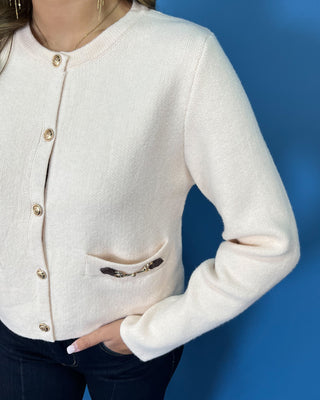 Ivory knit cardigan with gold buttons, long sleeves, front pockets, and decorative gold hardware.