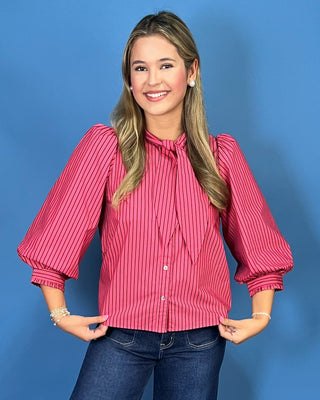 A woman modeling a pink and white vertically striped long-sleeve blouse with a tie-neck detail, paired with dark blue cropped jeans and silver shoes