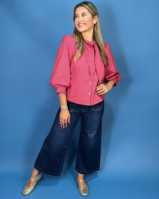 A woman modeling a pink and white vertically striped long-sleeve blouse with a tie-neck detail, paired with dark blue cropped jeans and silver shoes