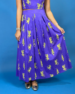 A model wearing a purple two-piece skirt set with yellow lion prints, standing against a solid blue background.