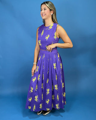A woman modeling a sleeveless purple maxi dress with a yellow and white lion print, standing against a solid blue studio backdrop.