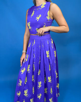 A woman wearing a sleeveless purple maxi dress with a matching waist tie and a full skirt, both decorated with yellow lion prints, set against a solid blue studio background.