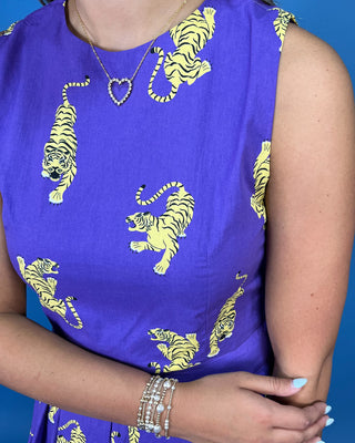A close-up of a sleeveless purple dress with a yellow and black tiger print, worn by a model with a silver heart necklace and stacked bracelets against a solid blue background.