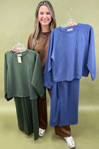 Woman holding a blue outfit with green and brown outfits displayed on hangers against a green background