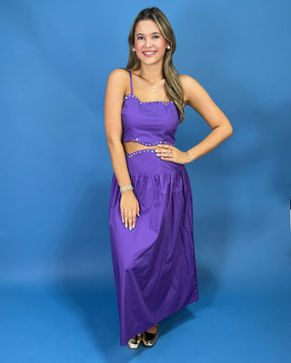 A woman modeling a purple two-piece maxi dress with spaghetti straps, a crop top, and a flared skirt, standing against a solid blue studio backdrop.