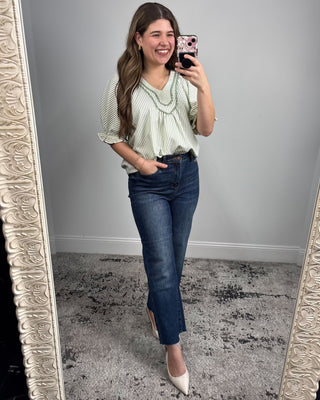 Woman taking a mirror selfie wearing a green blouse and blue jeans.