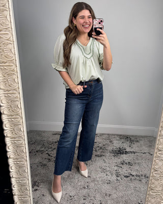 Woman taking a mirror selfie wearing a green blouse and blue jeans.