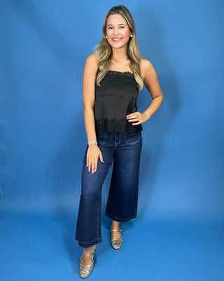 A woman modeling a black satin strapless top with lace trim and dark blue wide-leg jeans